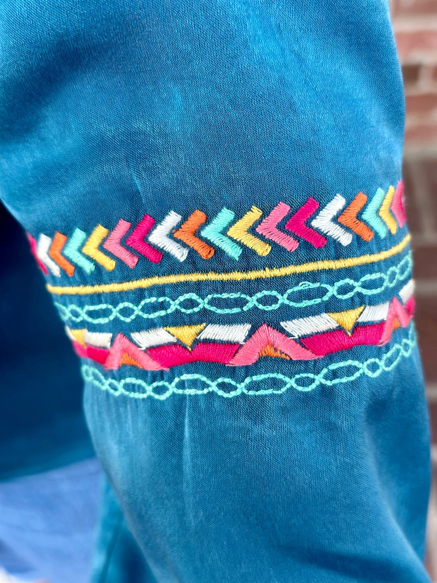 The Amy Top by Washco Apparel is an elegant teal shimmery fabric with colorful embroidery design around the v-notch ruffled neckline and sleeves. This top is long sleeve with an elastic band gathering around the wrist area, it can be pushed up or worn regularly.