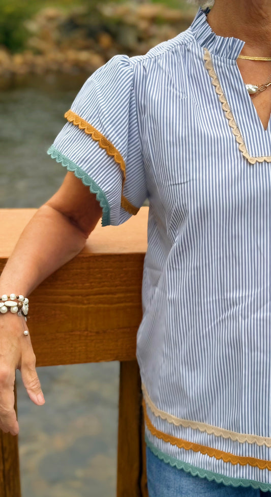 Woman wearing a light blue striped shirt with decorative trim, sitting by a body of water.