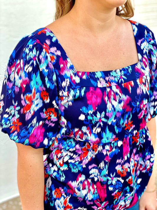 The Chandler Top by Washco Apparel is a bright, bold and multi-print baby doll top with puffed sleeves. The top includes a square neckline and smocking detail (with stretch) across the back.
