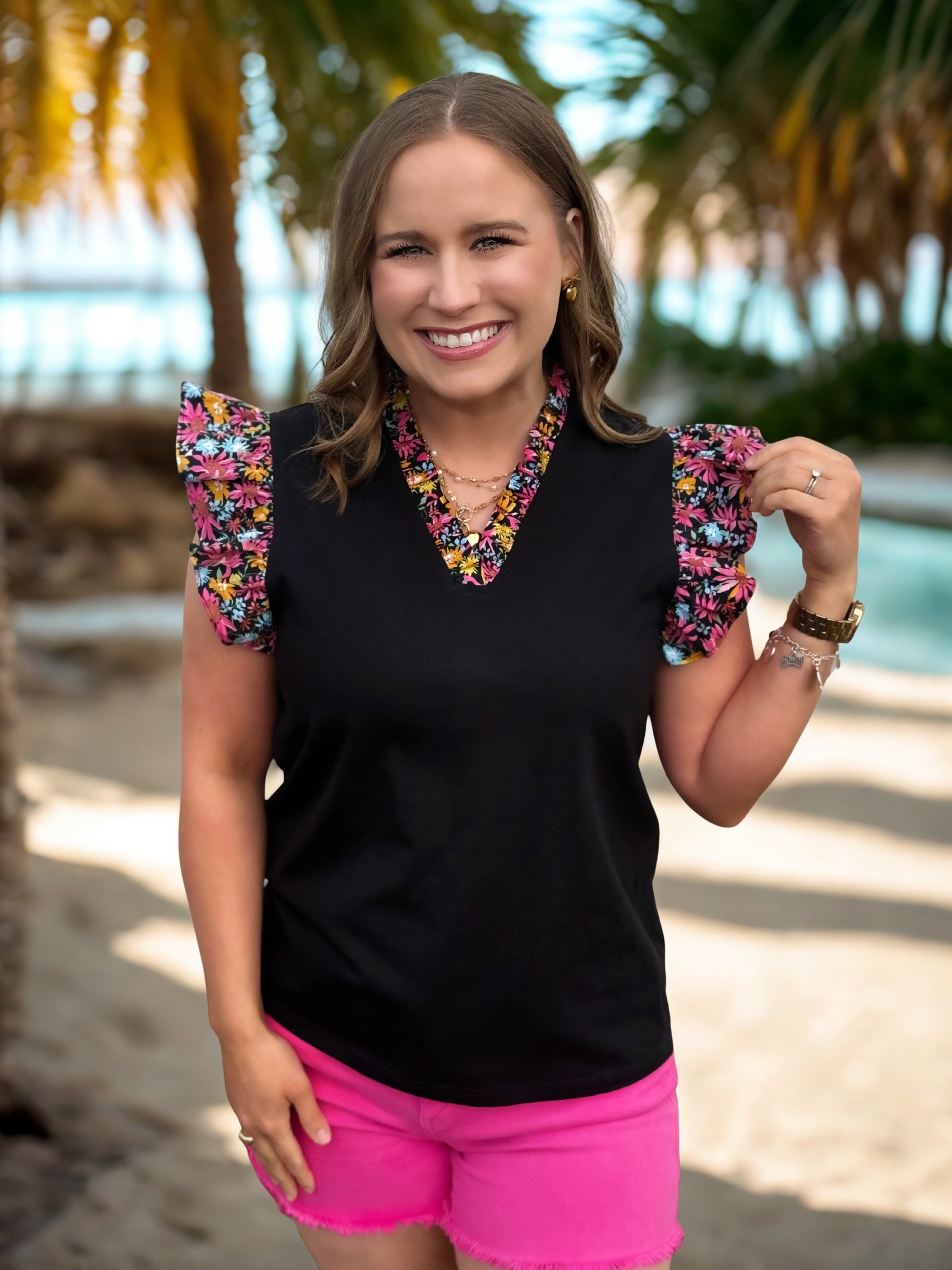 Woman wearing a black top with colorful sleeves and pink shorts, standing outdoors with palm trees in the background.