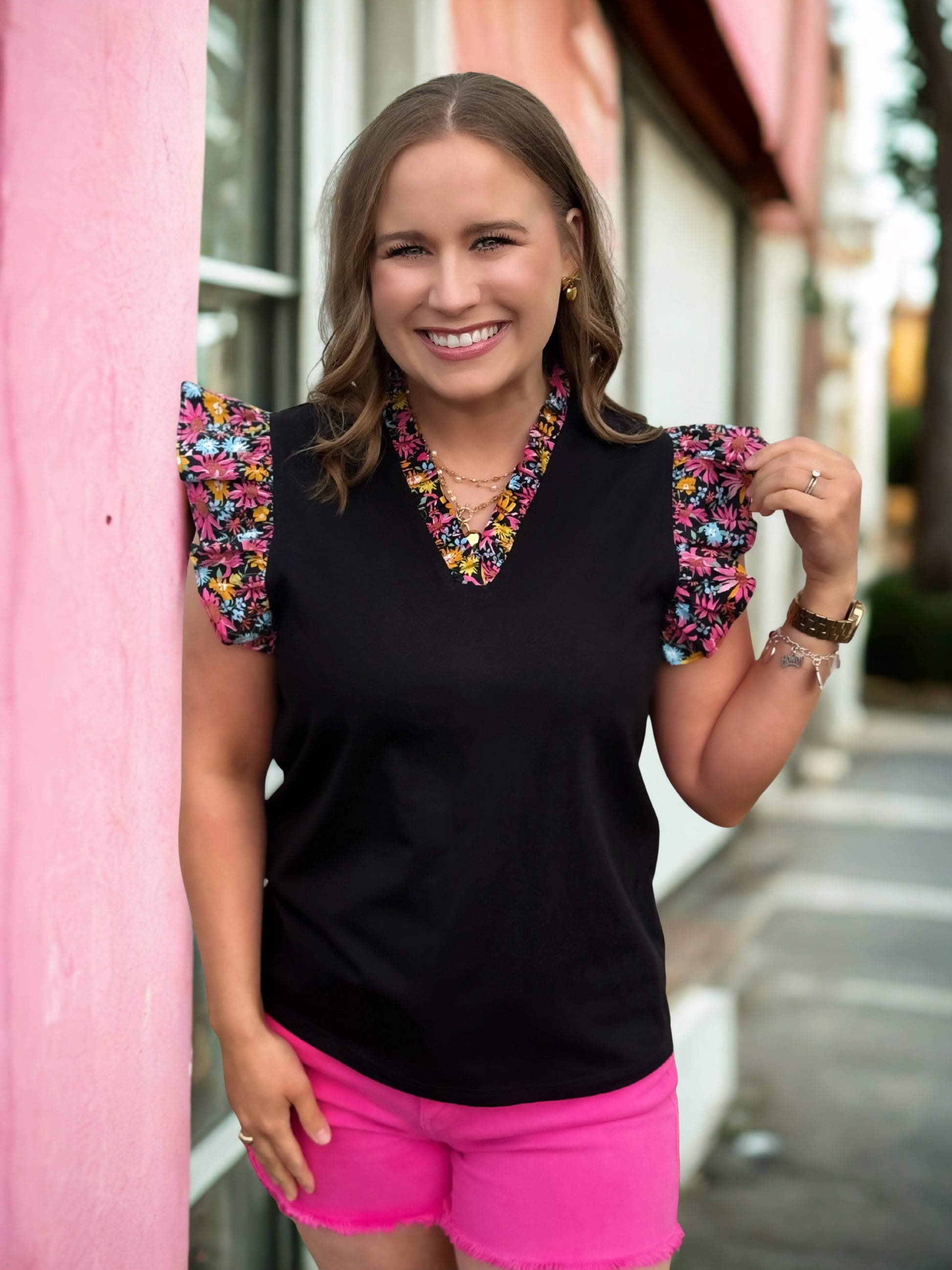 Woman wearing a black top with colorful sleeves and pink shorts, standing against a pink wall.